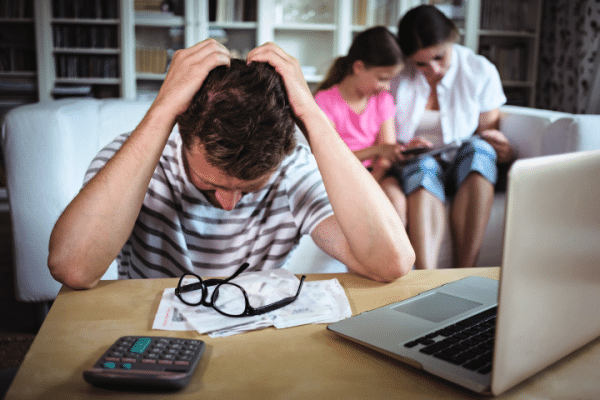 Man in striped t-shirt sits down at desk in front of documents and glasses and calculator and computer and he looks down in distress as he places hands over his head all while mom and daughter use tablet on white sofa behind him