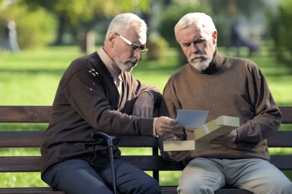 Two senior adults wearing brown sweaters and different pant colors while exchanging gift on park bench during sunny day
