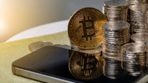 iphone on table next to gold bitcoin and stacks of crypto coins while photo has bright glimmer